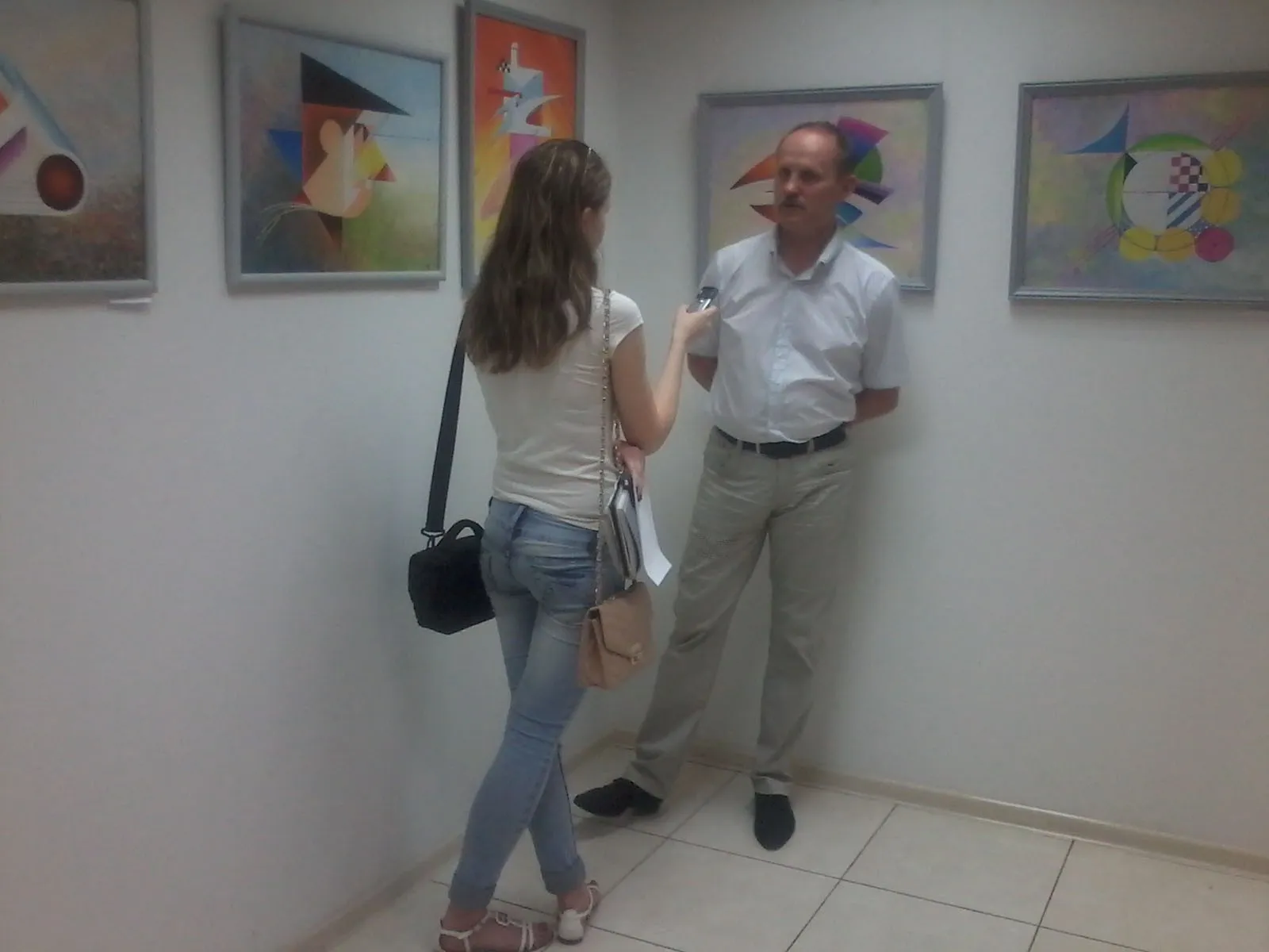 Oleksandr Zinoviev talking to a journalist in the gallery, his paintings on the wall behind him